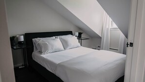 A neatly made bed in a room with a sloped ceiling, a bedside table with a lamp, and a small potted plant.