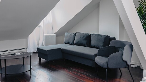A modern loft bedroom with a sloped ceiling, a gray sectional sofa, a round coffee table, and a potted plant.