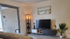 Ein Wohnzimmer mit Fernseher auf einem schwarzen Standfuß, einem Bild an der Wand, einer Pflanze und einem Sofa.