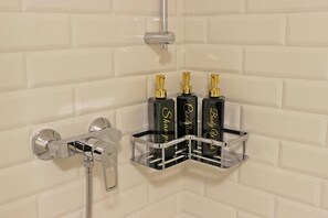 Ein Badezimmer mit weiß gefliesten Wänden, einer chromfarbenen Duschbrause und drei schwarzen Flaschen mit goldfarbenen Verschlusskappen in einem transparenten Behälter.