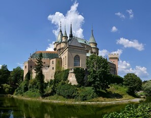 A historic castle with multiple towers, a moat, and lush greenery. - Galanda 24 (Turcianske Teplice)