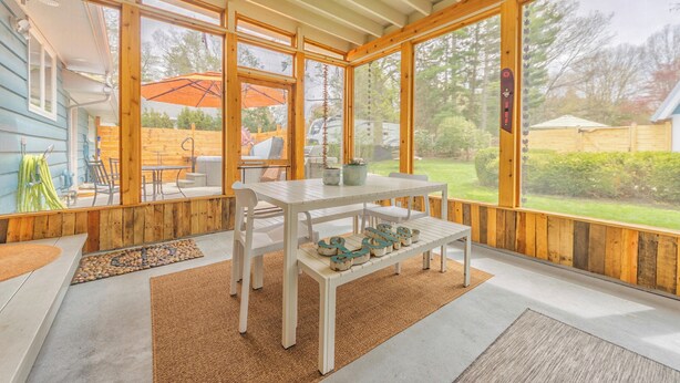 Un patio cubierto con una mesa blanca y bancos, ventanas amplias y vista al jardín trasero.