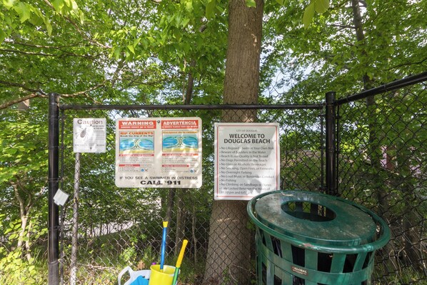Una cerca de malla con varios carteles de advertencia e información, un cubo de basura y un balde con utensilios de limpieza.