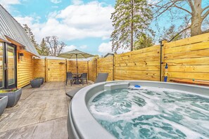 Jacuzzi en un área de terraza de madera con una mesa y sillas, rodeado por una valla de madera y un ventanal que da a un edificio.