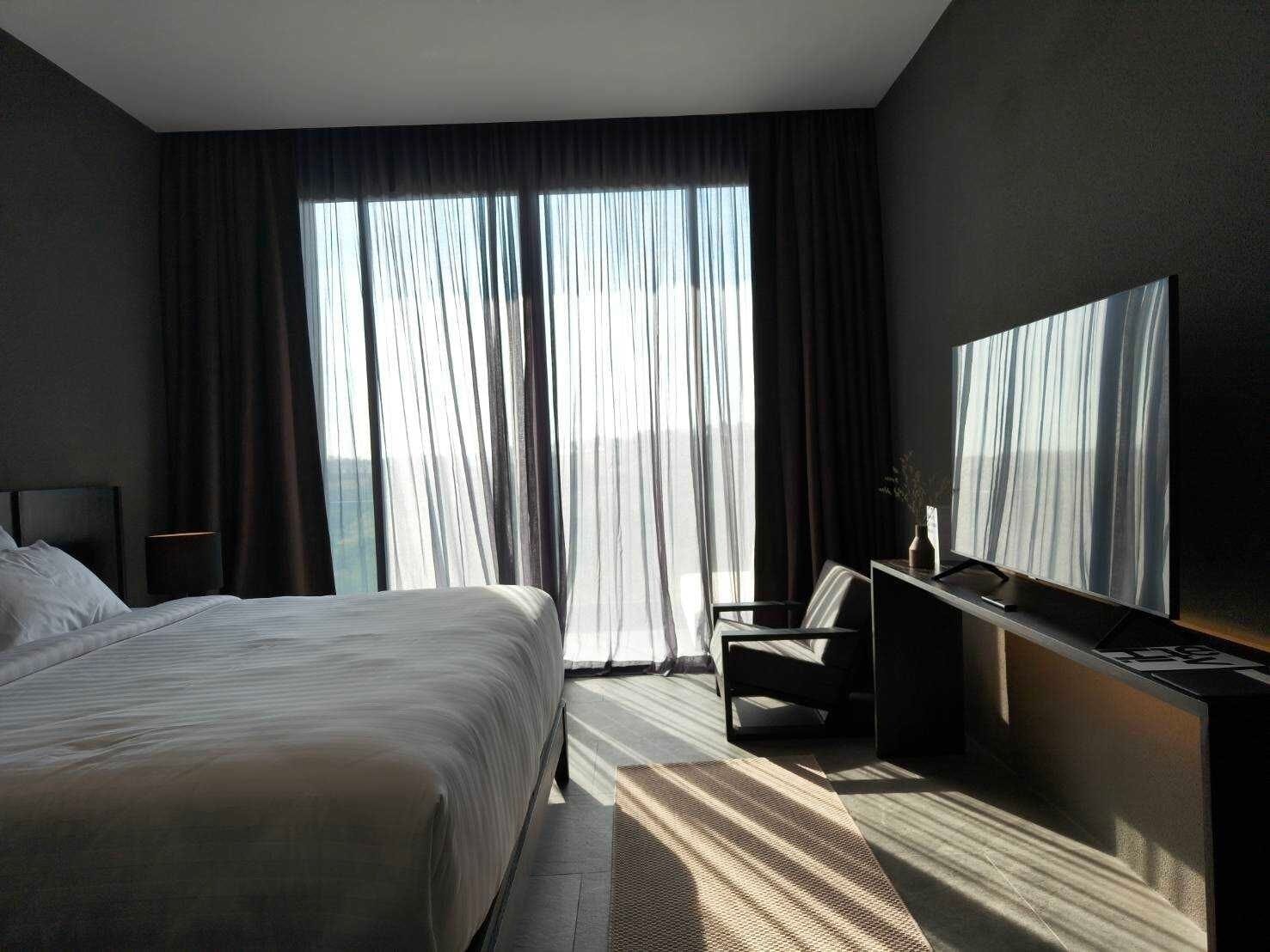 A modern hotel room with a large bed, a desk with a chair, a flat-screen TV, and a large window with curtains.