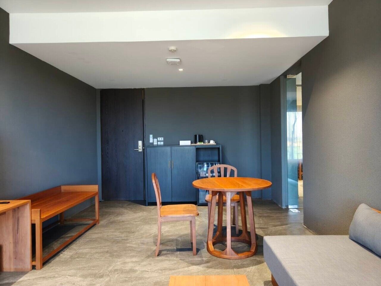 Un interior moderno con un banco de madera, una mesa redonda con dos sillas, un sofá y un mueble oscuro con refrigerador.