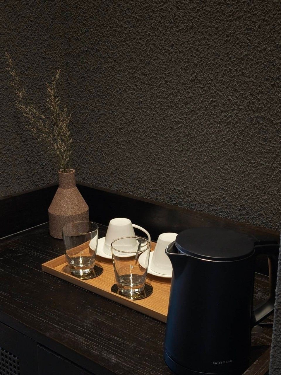 A tray with a water pitcher, glasses, and a vase with dried plants on a wooden table against a textured wall.