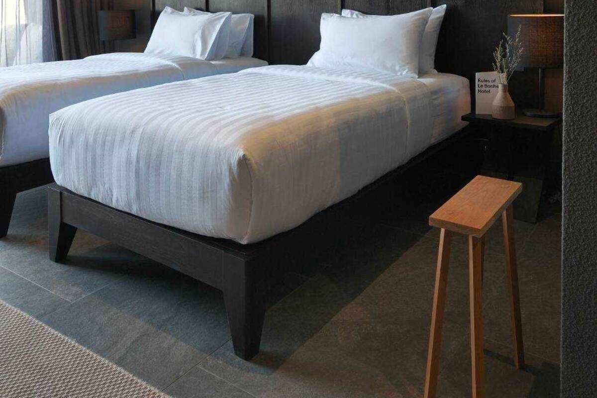 A hotel room with two beds, a small wooden stool, a nightstand with a lamp, and a sign indicating the hotel name.