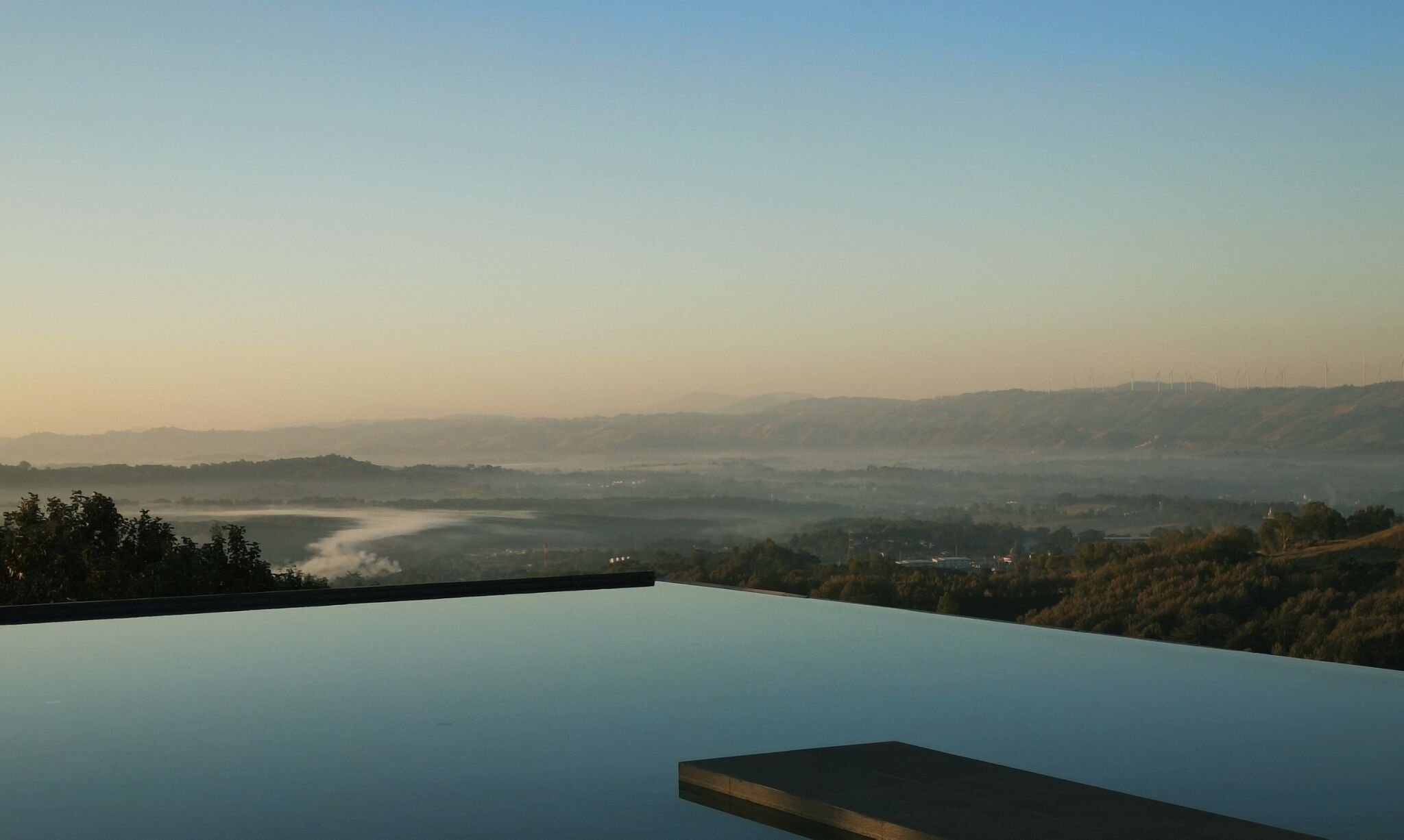 A panoramic view from a high vantage point overlooking a valley with a river, distant hills, and wind turbines.