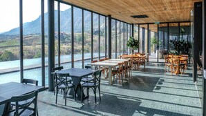 Een restaurant met grote ramen met uitzicht op een rivier en bergen, met houten tafels en stoelen.