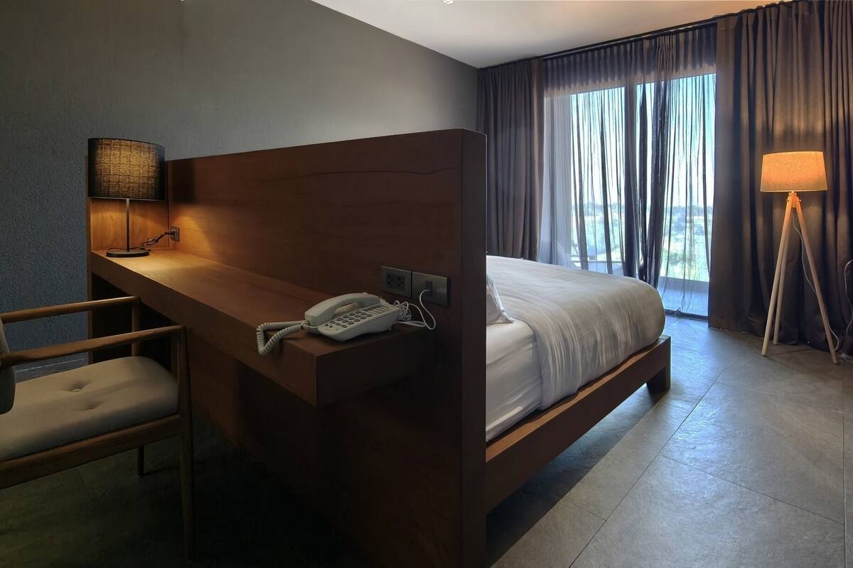 A hotel room with a wooden desk, a telephone, a chair, a bed, and a view of the outdoors through large windows.