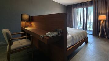 A hotel room with a wooden desk, a telephone, a chair, a bed, and a view of the outdoors through large windows.