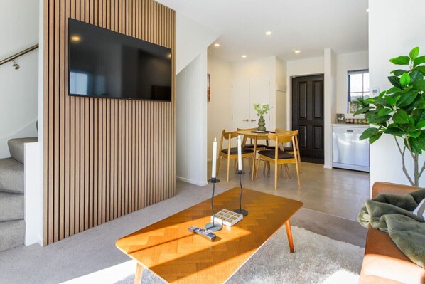 A modern living room with a wooden coffee table, a flat-screen TV mounted on a wooden panel, a dining area with a table and chairs, and a kitchen in the background.