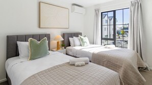 A hotel room with two beds, a bedside table with a lamp, a large window with curtains, and a wall-mounted air conditioning unit.