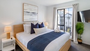 Una cama bien tendida con un edredón azul, almohadas blancas y un cabecero de madera. Hay dos mesitas de noche con lámparas, un ventanal con cortinas y una planta en maceta a la derecha.