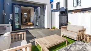 Un patio exterior con mobiliario de madera, puerta de vidrio que da a un interior moderno y un pequeño jardín.