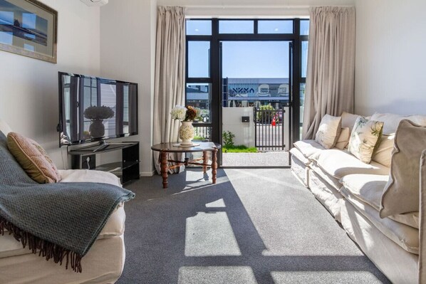 A living room with a sofa, a TV, a small table with a vase, and a view of a gate and greenery outside.