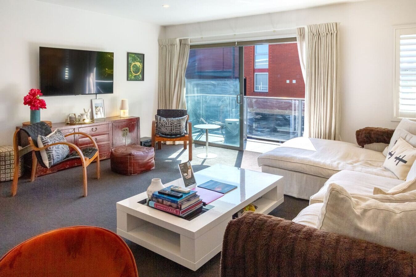 Una sala de estar moderna con sofá, sillón y mesa de centro. Hay un televisor de pantalla plana montado en la pared y una ventana con cortinas.