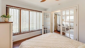 Un dormitorio con cama, ventilador de techo, ventana con persianas y puerta que da a otra habitación.