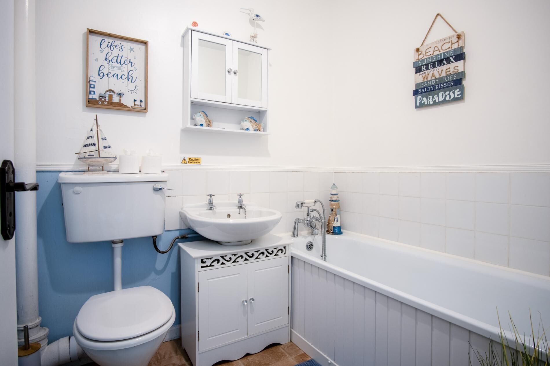 A bathroom with a toilet, sink, and bathtub. There is a framed sign on the wall and a shelf with toiletries.