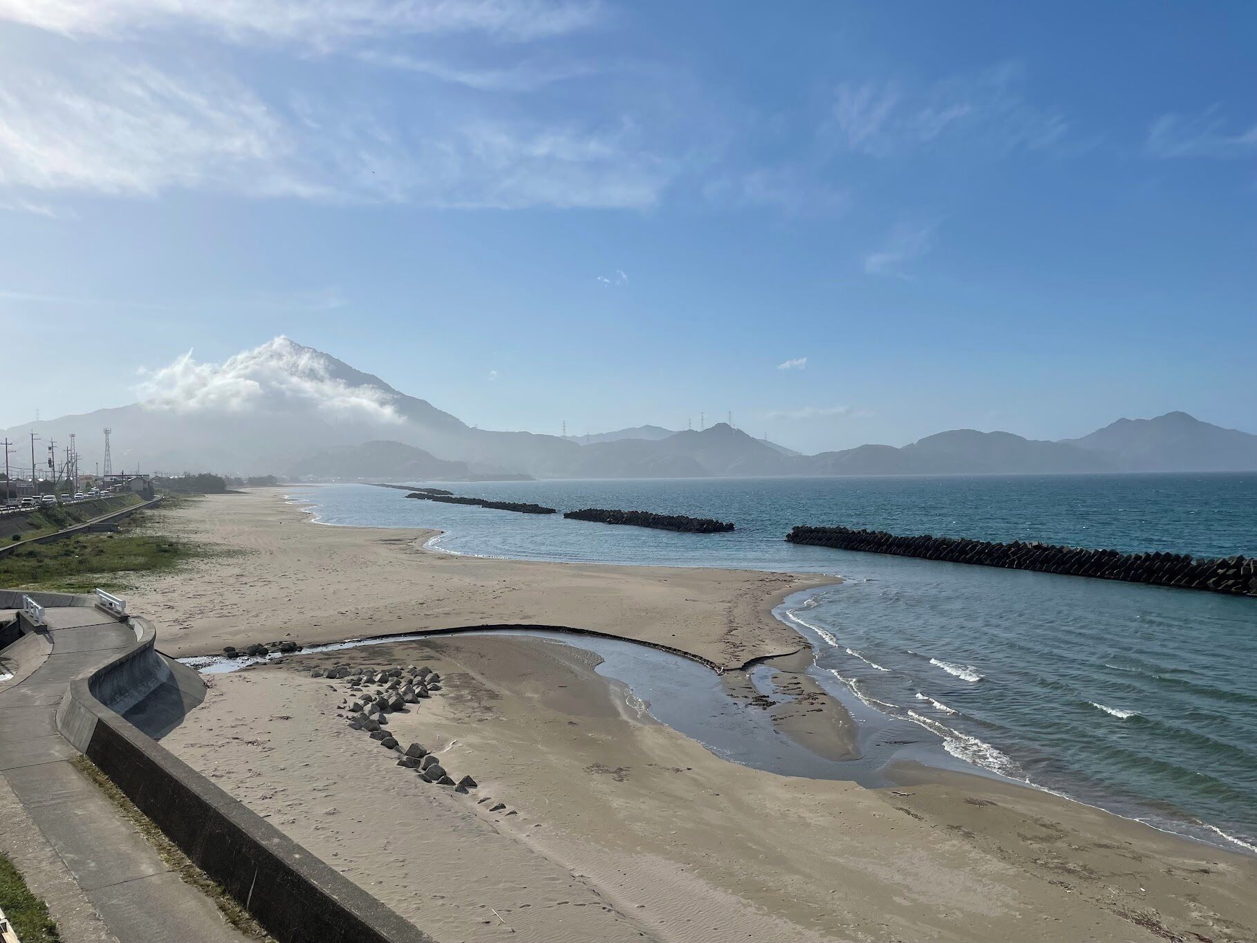 A coastal beach with a clear sky, a mountain in the background, and a road parallel to the beach.