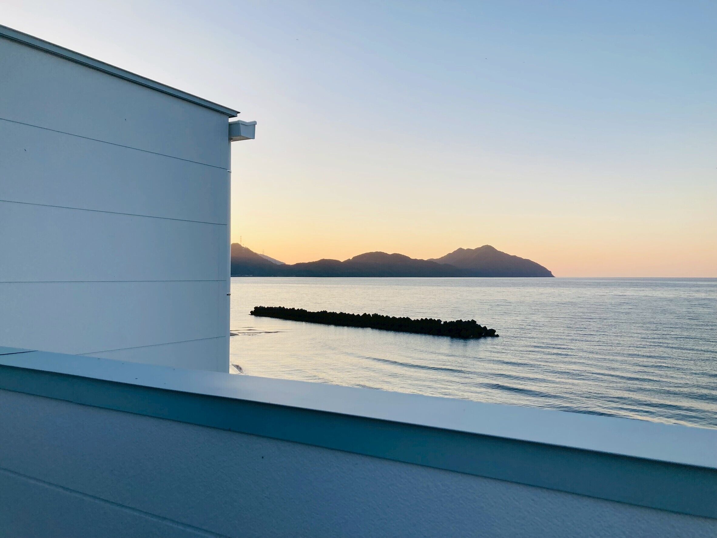 A view of a calm sea with a breakwater and distant mountains during sunset.