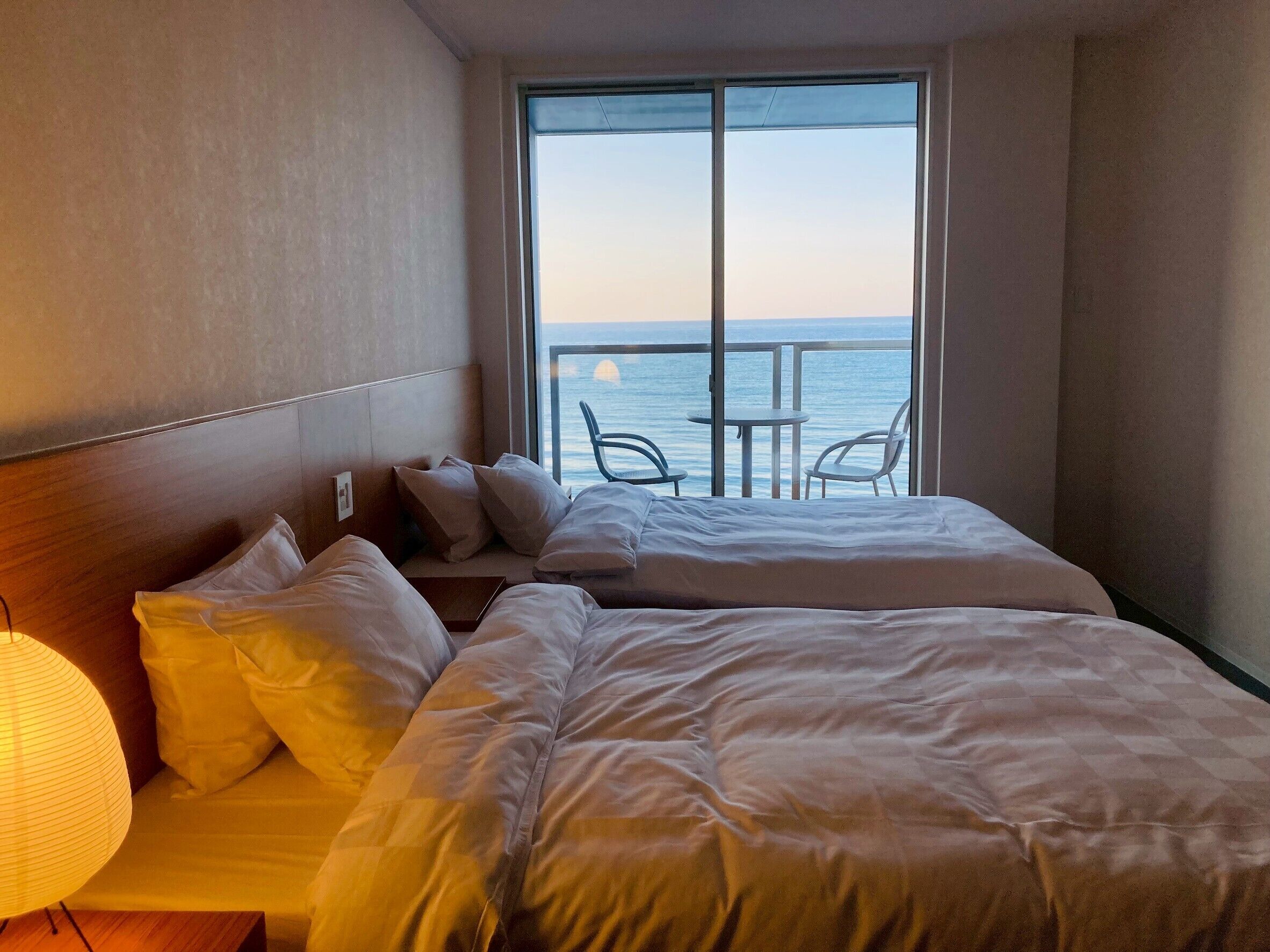 A hotel room with two beds, a nightstand, a lamp, and a view of the sea through large windows.