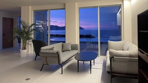 Ein modernes Wohnzimmer mit Sofa, Sessel und Couchtisch, große Schiebeglasfenster, die auf einen Balkon mit Blick auf das Meer und die Berge führen.