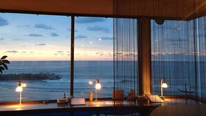 Ein Zimmer mit großem Fenster mit Blick auf das Meer bei Sonnenuntergang. Es gibt drei Tische mit Lampen und Stühlen.