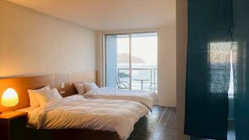 A hotel room with two beds, a nightstand with a lamp, a large window with a view of the sea, and a balcony with chairs.