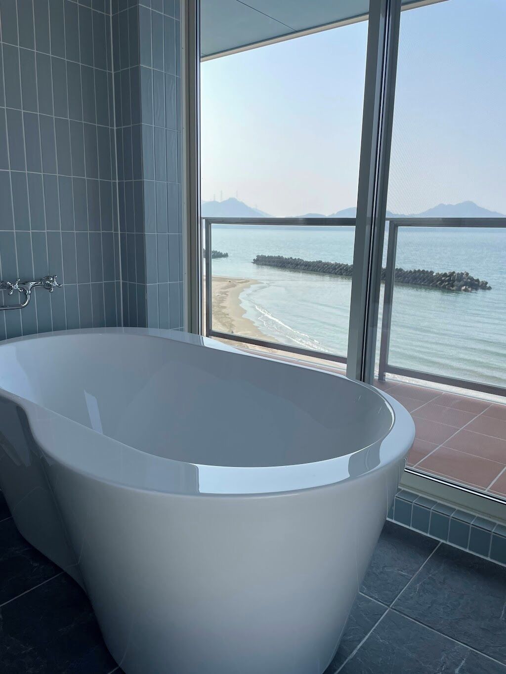 A modern bathroom with a freestanding bathtub, large windows offering a view of the sea and distant mountains.