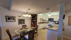 A modern kitchen with white cabinets, a microwave, and a stove. There is a dining area with a glass table and wicker chairs. - CP 413 Dune View Condo - Welcome To Paradise! (Fort Pierce)