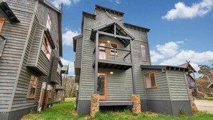 A two-story wooden cabin with a porch and stone pillars, surrounded by grass and trees. - The Chalet (Dinner Plain)