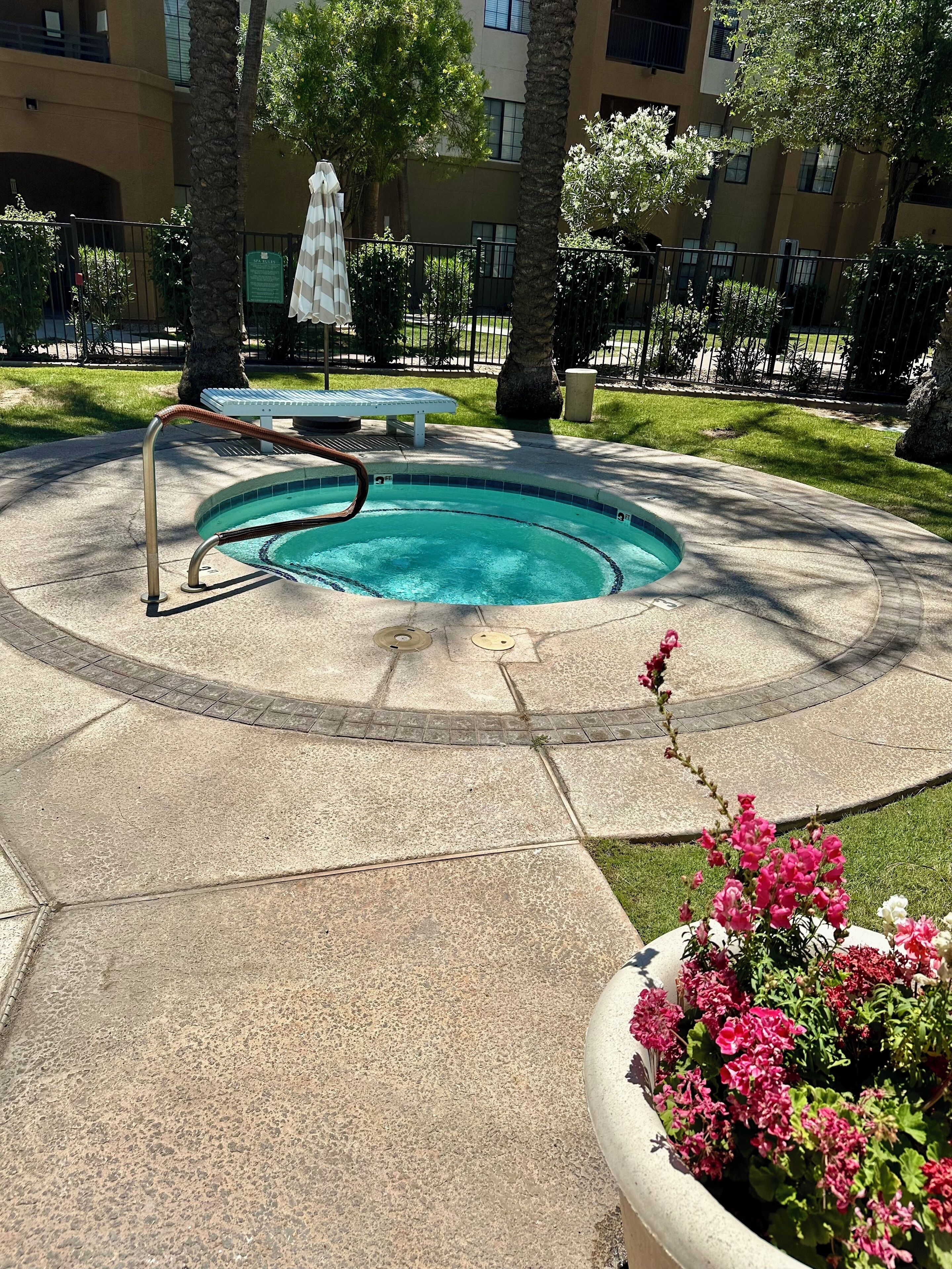 A swimming pool with a metal ladder, surrounded by a concrete deck. There is a bench and a small umbrella in the background.