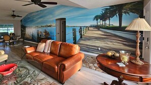 Un salon avec une grande fresque murale représentant une plage et des palmiers, un canapé en cuir marron, une table ronde en bois avec une lampe, et une table basse en verre.