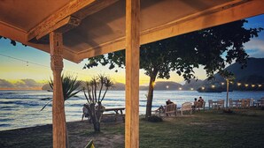 Een terras buiten met houten meubels en tafels, met uitzicht op een waterpartij en een bergachtig landschap.