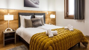 A bedroom with a wooden headboard, two bedside lamps, a bed with white bedding and a mustard-colored quilt, a painting above the bed, and a window with curtains. - Apartment in Plagne 1800 Near Paradiski (La Plagne-Tarentaise)