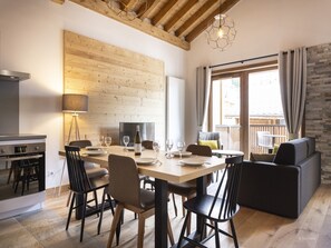 Zona pranzo con tavolo in legno, sedie e divano. C'è un angolo cottura con piano cottura e lampada. La stanza presenta travi a vista al soffitto e una finestra con tende.