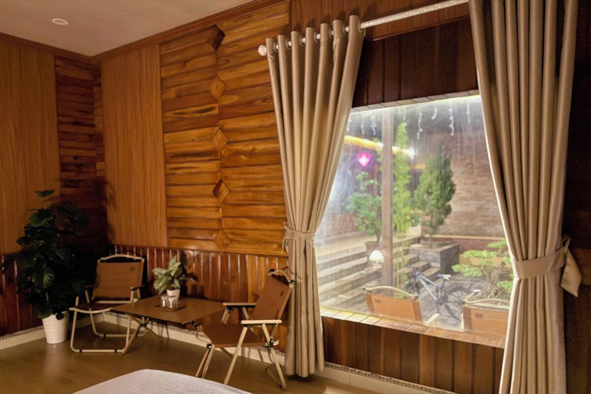 Ein Zimmer mit Holzwand, einem Vorhangfenster, einem Bett, einem Tisch mit Stühlen und einer Topfpflanze.