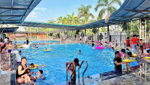 A swimming pool area with a covered deck, palm trees, and people swimming and relaxing. - May Garden Homestay (Ninh Binh)