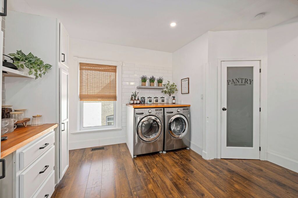 A laundry room with two front-loading washing machines, a wooden countertop with jars, and a window with blinds.
