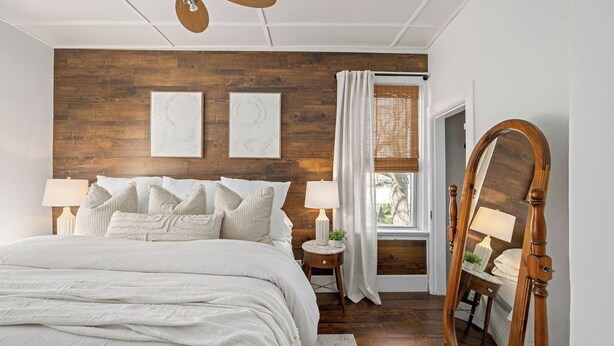 Ein Schlafzimmer mit einer Wand in Holzoptik, einem großen Bett mit weißer Bettwäsche, einem stehenden Spiegel und zwei Nachttischlampen.