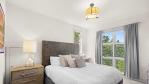 A bedroom with a large bed, a nightstand with drawers, a lamp, and a window with curtains.