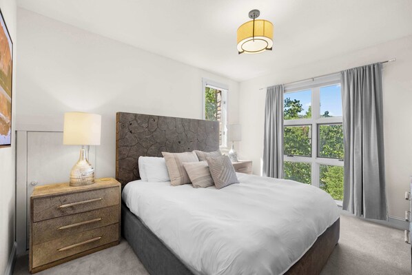 A bedroom with a large bed, a nightstand with drawers, a lamp, and a window with curtains.