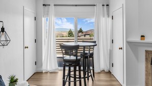 A room with a wooden floor, a round table with chairs, a window with curtains, and a view of a landscape.