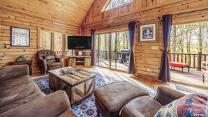 Un intérieur de chalet chaleureux avec des murs en bois, un grand téléviseur, des sièges confortables et une terrasse offrant une vue magnifique.