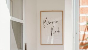 Een lijst met de naam 'Barrie Noo' op een witte muur, een houten deur en een kleine ruimte met een tafel en een plantje.