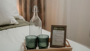 Een bed met een dienblad waarop een glazen fles, twee groene kandelaars en een menukaart in een lijstje staan in een hotelkamer.