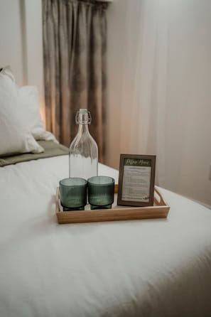 Une table de chevet sur laquelle se trouve un plateau avec une bouteille en verre, deux bougeoirs verts et un menu encadré, dans une chambre d’hôtel.