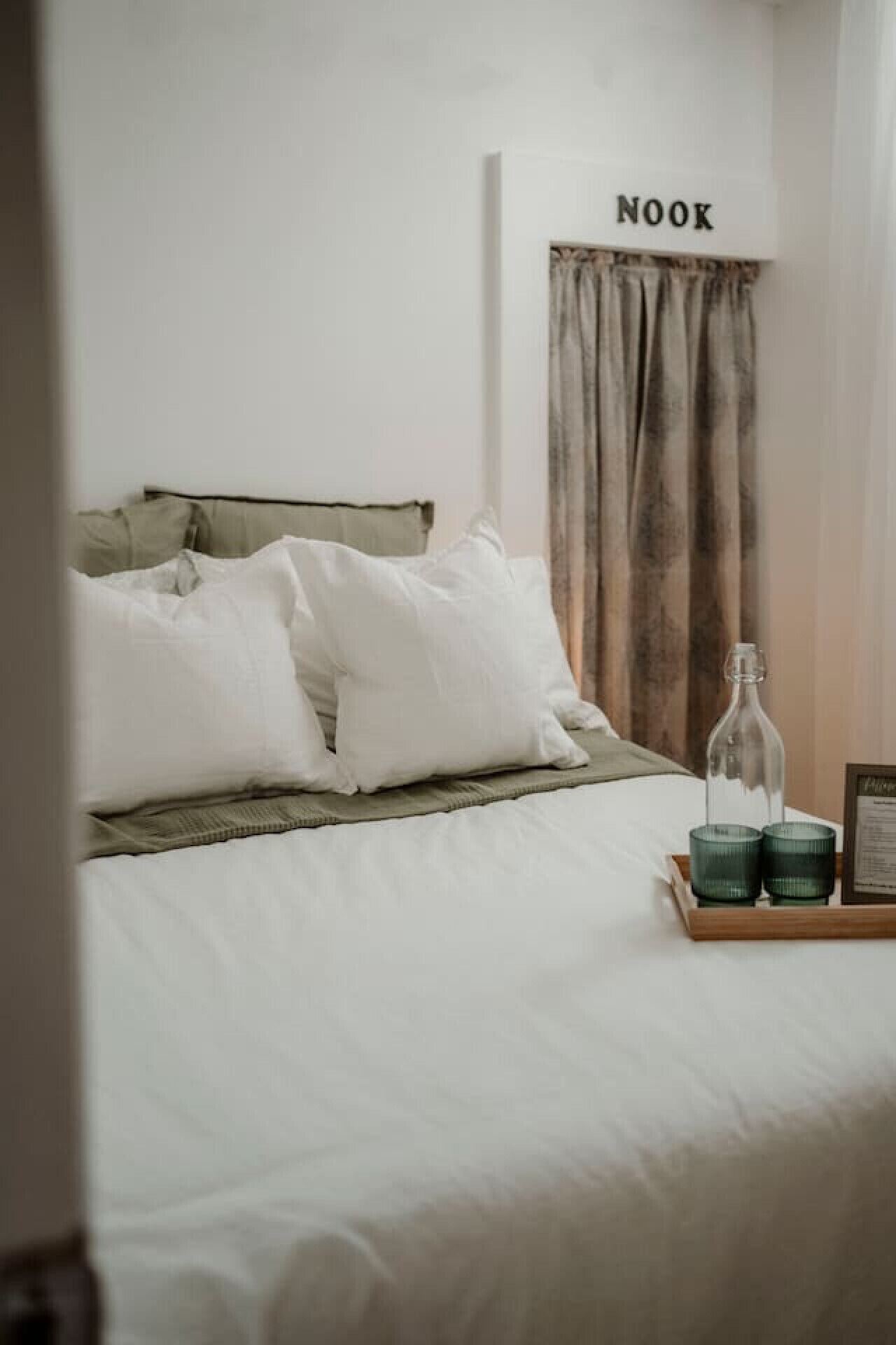 Una cama bien hecha con ropa blanca y una manta verde, una mesita de noche con una botella y vasos, y un letrero sobre la cama que dice 'NOOK'.
